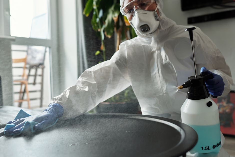Person in protective gear disinfecting a table indoors, ensuring safety amid COVID-19.